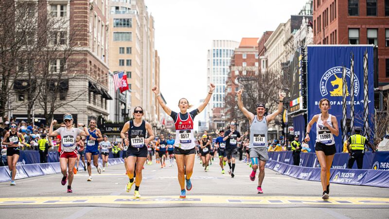 The image shows a marathon race with several runners approaching the finish line. The runners are in motion, with their arms raised in celebration. The background includes buildings and spectators, suggesting a large event. The atmosphere seems energetic and celebratory.